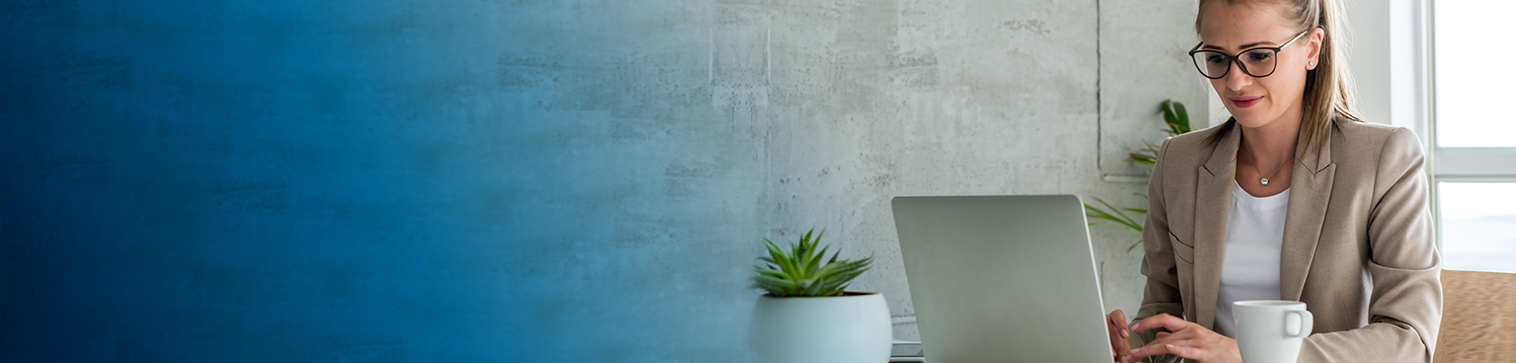 Imagen mujer con gafas y blazer viendo un computador en un escritorio con una taza de café y una planta